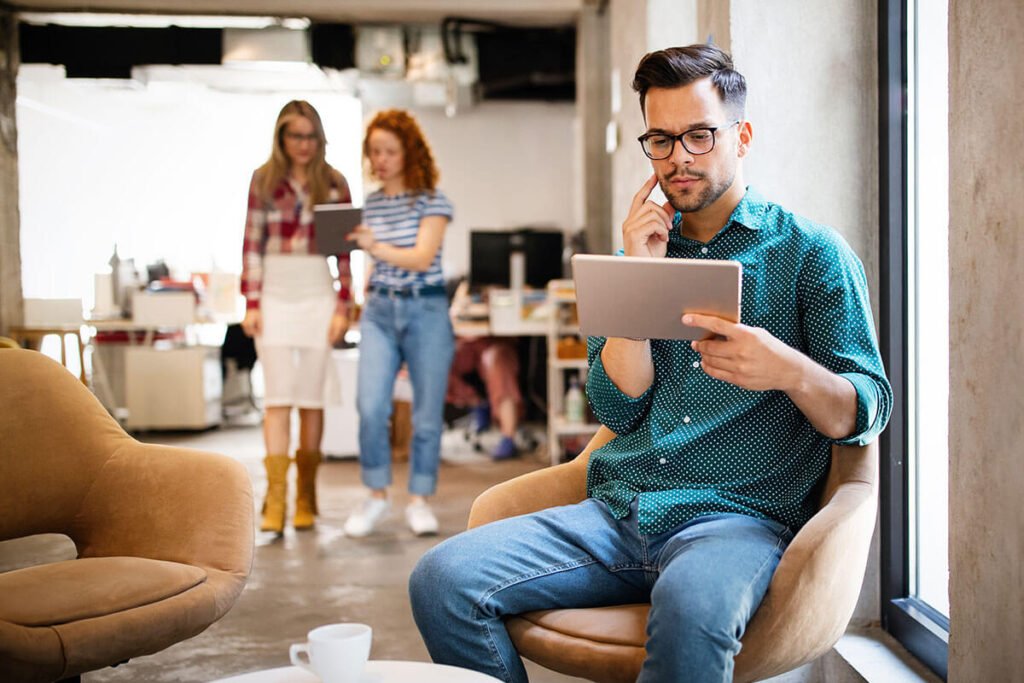 Man reviewing ideas on a tablet in a modern office while teammates discuss projects in the background.